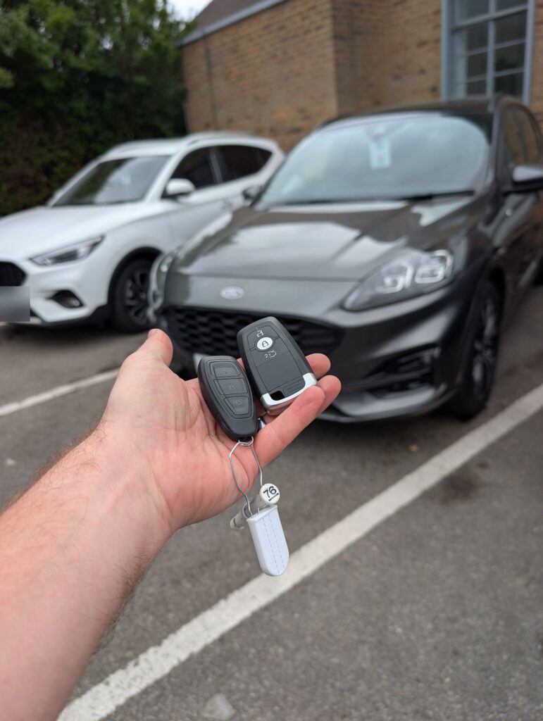 Two Ford keyless fobs held in front of a grey Ford Kuga in Sittingbourne